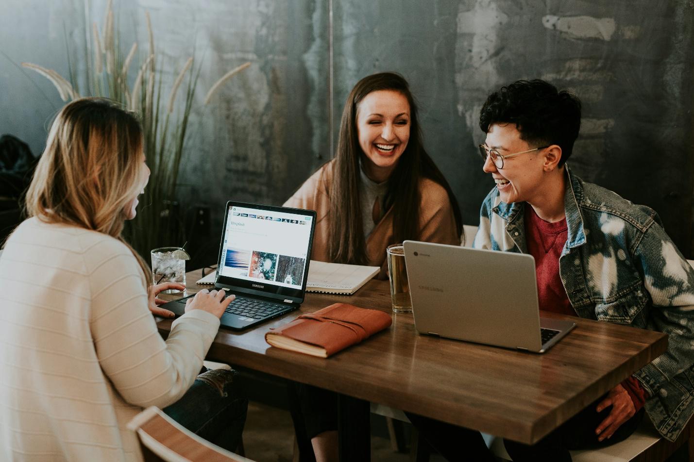 Three employees sitting on a desk with laptops, having a healthy conversation and laughing