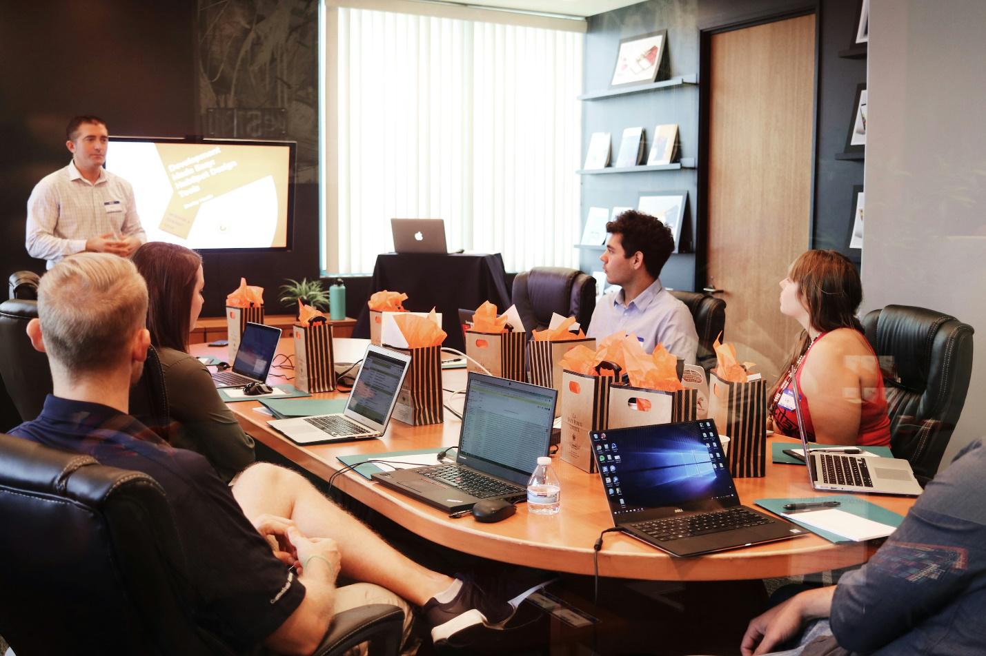 A man giving training to employees who are sitting at a round table with their laptops