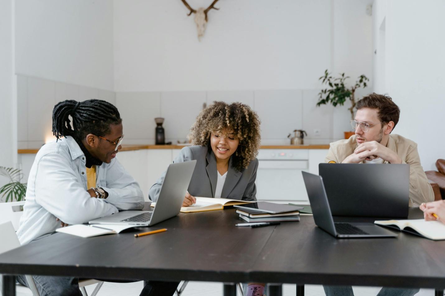 A picture of three employees sitting together with laptops, diaries and pens on the table